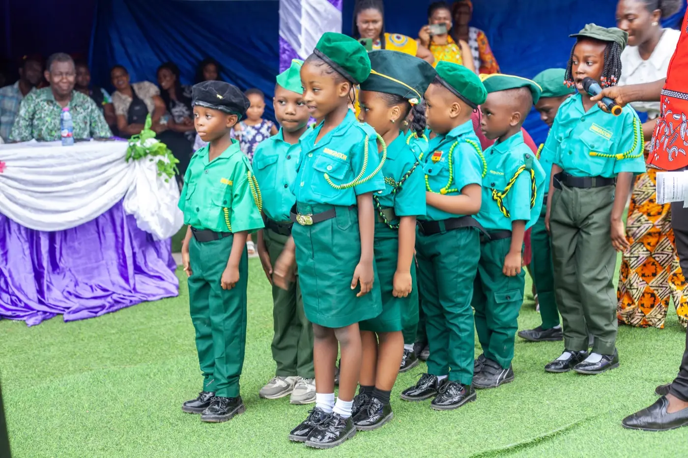 Preschool pupils in green ceremonial uniforms and berets lined up to perform at a school event
